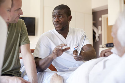 Male nurse discussing over digital tablet with couple while senior man reclining at hospital ward