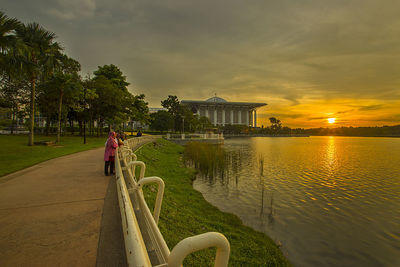 People standing by lake against sky during sunset