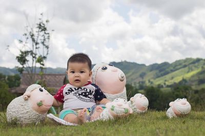 Portrait of cute baby boy sitting with sheep toys on field against sky