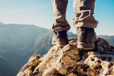 Unrecognizable crop explorer in trekking boots standing on rocky hill in highlands on sunny day