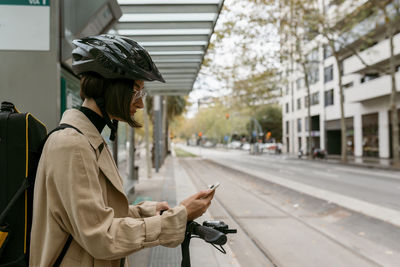 Mid adult woman with instrument case and electric push scooter using mobile phone while standing at tram station in city