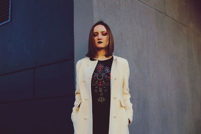 Portrait of young woman in white coat standing against wall