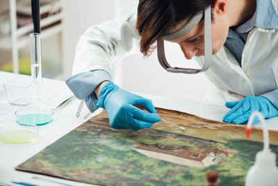 A female conservator with magnifying goggles repairing damages on an oil painting canvas.