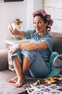 Woman with hair curlers taking selfie while sitting on sofa at home