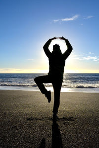 Silhouette person practicing yoga while standing on sea shore against sky during sunset