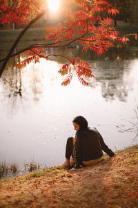 Young woman sitting by lake under a tree during autumn