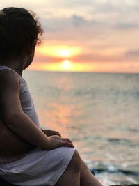 Midsection of man sitting at beach against sky during sunset