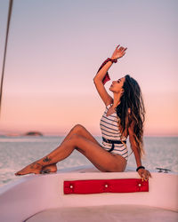 Woman with arm raised sitting in nautical vessel on sea against sky during sunset