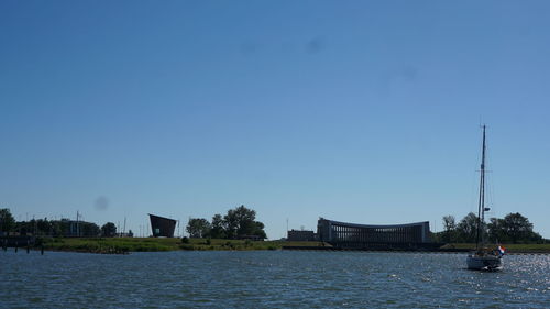 Sailboats in river by buildings against clear blue sky