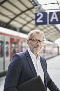 Senior businessman with laptop standing on railroad station platform