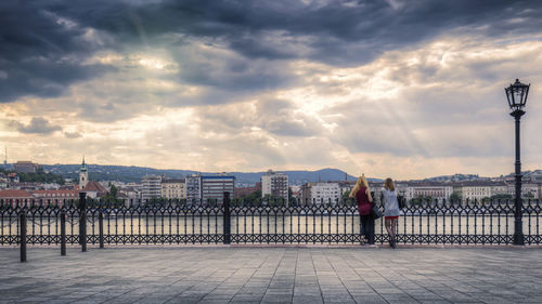Rear view of women standing on promenade in city against cloudy sky