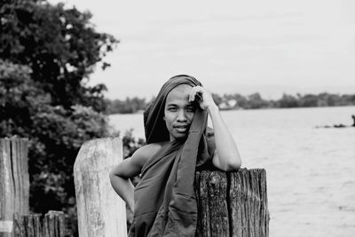 Portrait of monk leaning on wooden post while standing against lake