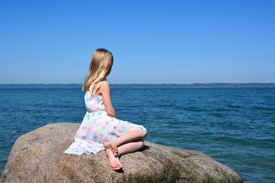 Side view of girl sitting on rock by sea against clear sky