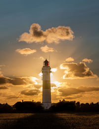 Lighthouse on field by building against sky during sunset