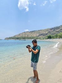 Full length of man holding camera standing on beach against sky