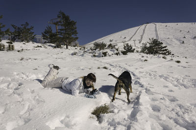 Portrait of smiling woman with dog relaxing on snow covered landscape against clear blue sky