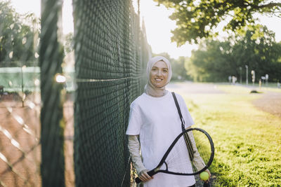 Portrait of smiling woman holding tennis racket near fence
