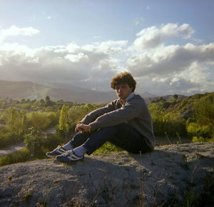 Side view of young man sitting on mountain against sky