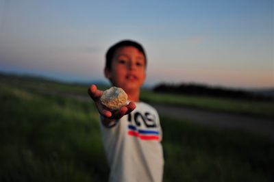 Boy holding stone on field against sky during sunset