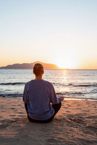 Rear view of woman sitting in yoga poses at beach against sky during sunset