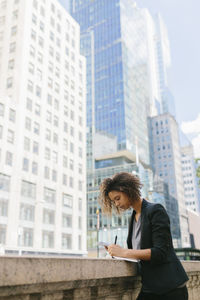 Afro female professional writing while leaning on retaining wall near office buildings