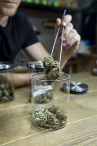 Crop anonymous male with tweezers taking out dry cannabis flower buds of jar on table in workspace