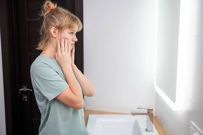 A teenage girl with problematic skin is getting ready to wash in the bathroom and holding her face