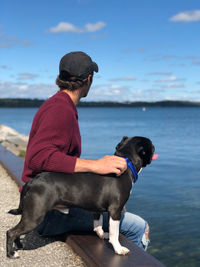 Man with dog sitting by lake against sky