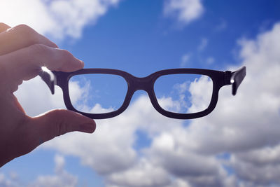 Close-up of hand holding eyeglasses against sky