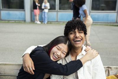 Cheerful teenage girl embracing male friend while sitting on bench