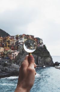 Cropped hand holding magnifying glass against buildings on mountain by sea