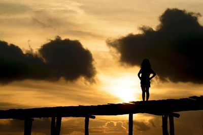 Low angle view of silhouette girl standing on jetty against sunset sky