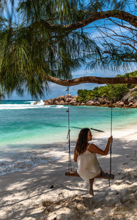 Rear view of young woman wearing beach clothes, sitting on swing on paradise beach