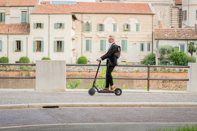 Side view full length young businessman in a suit riding an electric scooter while commuting to work