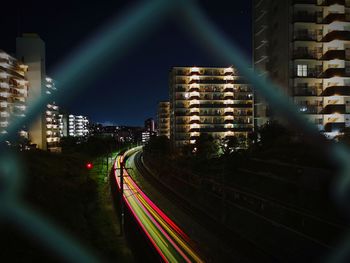 Light trails on road amidst buildings seen through chainlink fence