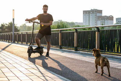 Handsome man is standing one leg on electric scooter and looking on his dog on the street. 