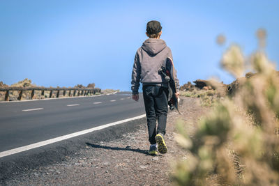 Rear view full length of boy walking with skateboard at roadside