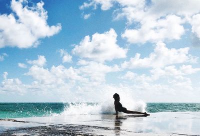 Water splashing on silhouette woman posing at beach against cloudy sky