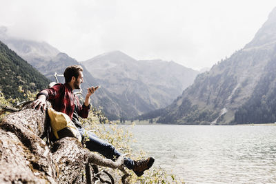 Austria, tyrol, alps, hiker on tree trunk at mountain lake using cell phone