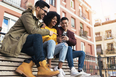 Smiling young woman with friends using phones while sitting on bench