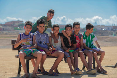 Portrait of friends sitting on bench against sky