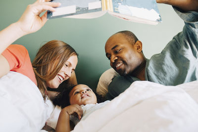 Happy parents showing storybook to daughter while sleeping together on bed at home