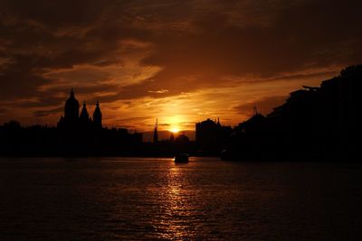 Silhouette buildings by river against sky during sunset