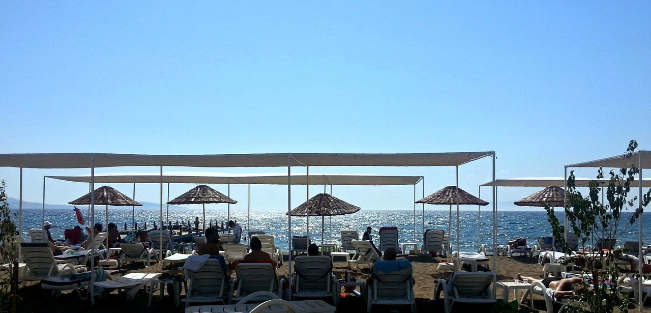 sea, clear sky, beach, chair, water, copy space, horizon over water, parasol, beach umbrella, large group of people, sunshade, vacations, lounge chair, restaurant, table, blue, shore, sand, relaxation, sunlight