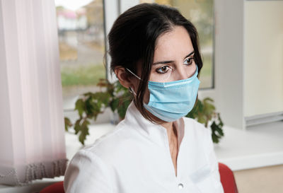 Tired exhausted woman nursewearing medical unform and face mask sitting in front of clinic window 