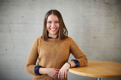 Portrait of smiling female student standing by wooden table against wall in university