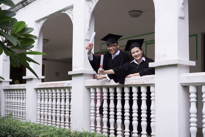 Portrait of smiling students wearing graduation gowns while leaning on railing