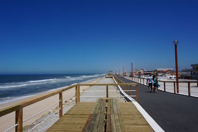 People walking on pier at beach against clear blue sky