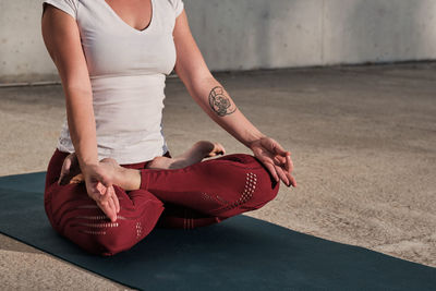 Cropped unrecognizable barefooted woman with tattoo in white shirt and burgundy pants sitting in padmasana with gyan mudra and meditating against concrete wall on street among urban environment