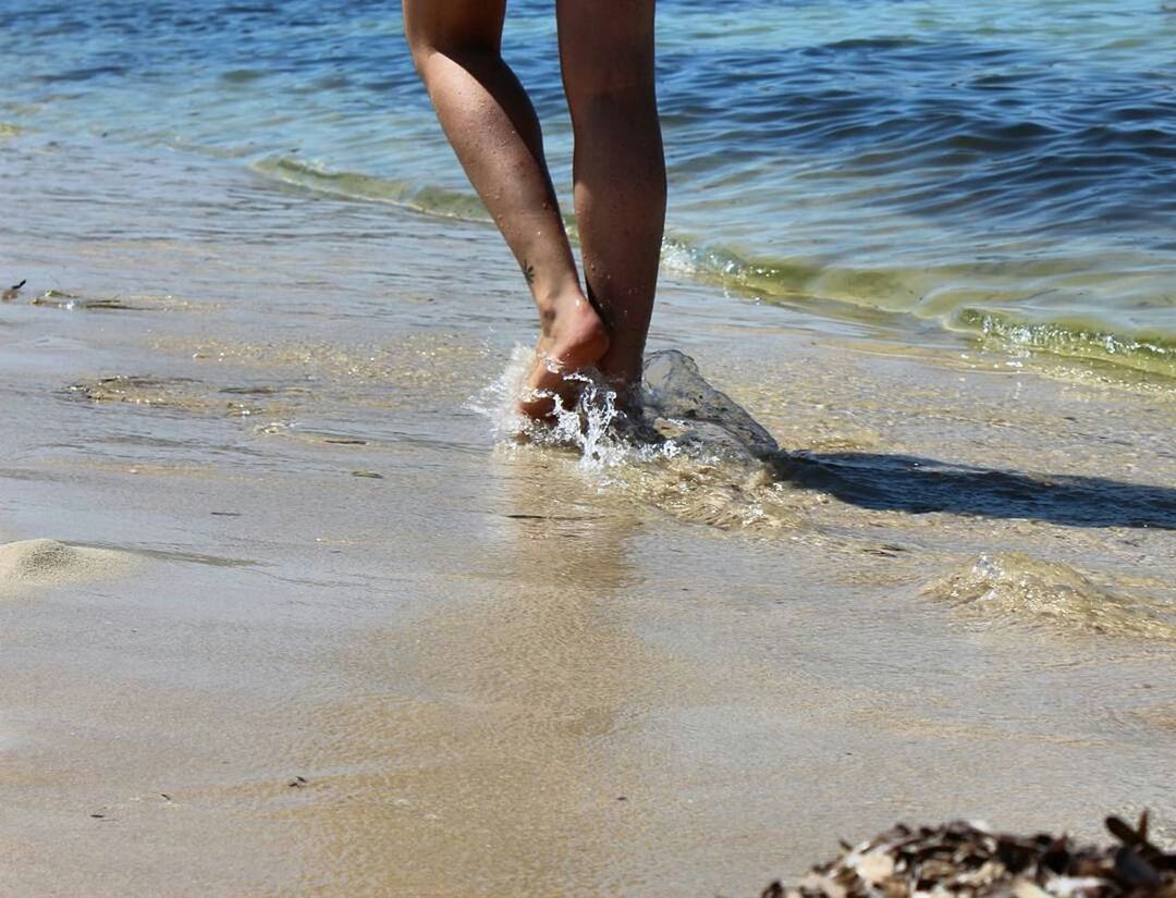 low section, person, water, beach, barefoot, lifestyles, sand, leisure activity, standing, sea, lake, vacations, wet, human foot, shore, outdoors, day, wave, nature, tranquil scene, tranquility, tourism, escapism